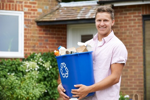 A young man with short, light brown hair and a bright smile stands outdoors in front of a brick house with a white-framed window and a sloped roof. He is wearing a light pink, short-sleeved shirt and is holding a large blue rubbish bin with a white recycling symbol on the side. The bin is filled with a variety of waste materials, including white and brown paper, plastic bottles, and other household rubbish. In the background, there are green shrubs and flowering plants along the side of the house, suggesting a residential garden environment. The scene is well-lit with natural daylight, indicating a clear day. This image is relevant to rubbish removal services offered by Garden Clearance Shepherds Bush, highlighting the act of collecting household waste for disposal or recycling in a tidy, suburban setting in Shepherds Bush, London.