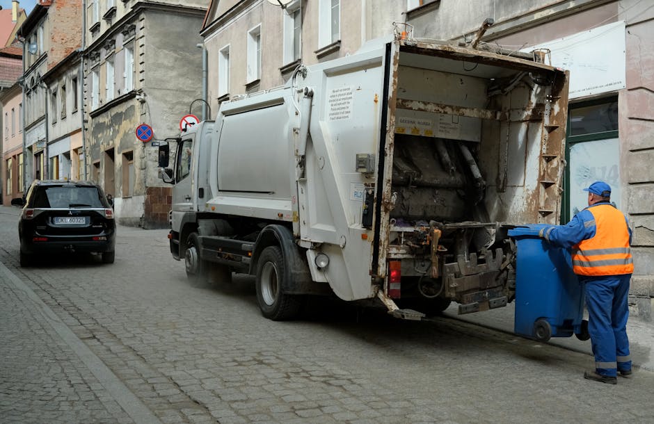 A street scene featuring a white bulk collection vehicle parked next to a building with aged, weathered facades and multiple windows. The rear of the vehicle is open and loaded with mixed waste materi